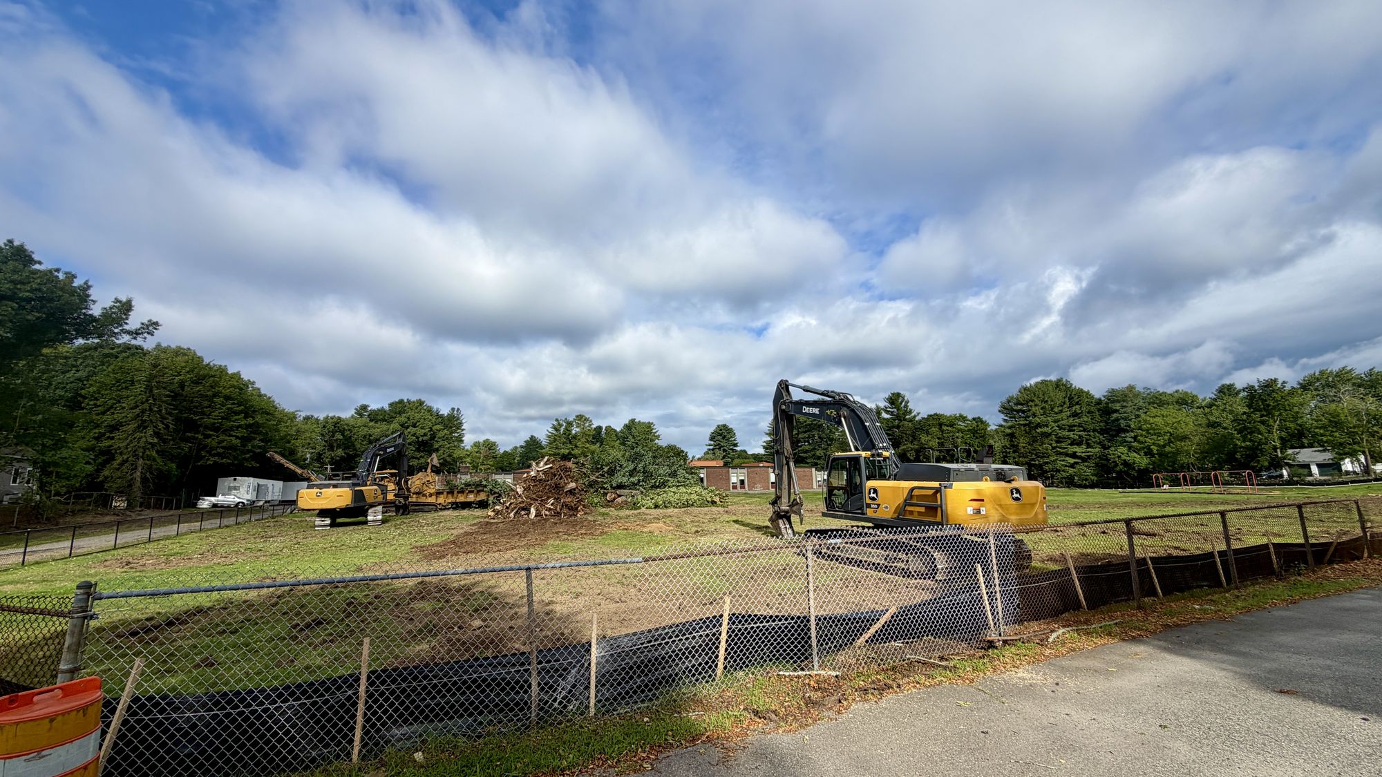 Killam site, photographed August 6, 2026: two excavators clear the field behind chain-link construction fencing, a pile of tree stumps and brush at the center, with the 1969 school building and playground visible in the distance under a partly cloudy sky.
